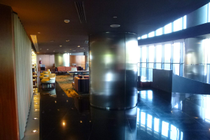 Eine geräumige Business-Lounge am Hong Kong International Airport mit großen Fenstern, hohen Decken mit elektrischen Leuchten, Sitzmöglichkeiten wie Stühle und Sofas, Tischen, einem Teppichboden und einer zentralen Säule.