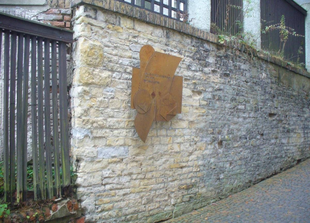 Steinmauer mit Metalltor auf der linken Seite, eine Tafel, die die Eröffnung des alten Rathauses erinnert, und ein Gebäude mit Fenstern und Pflanzen im Hintergrund.