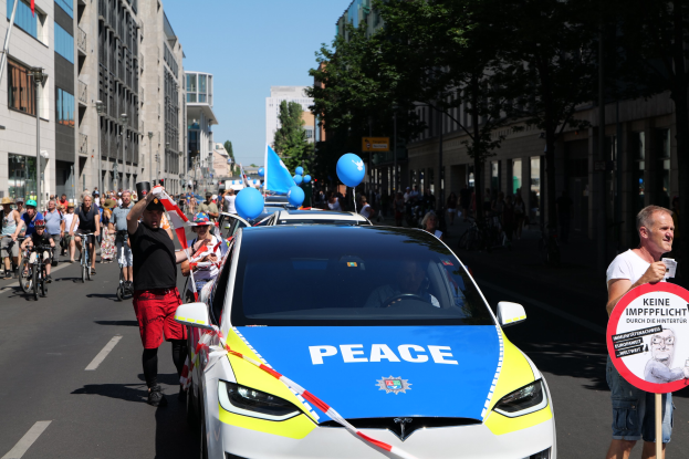 Eine Gruppe von Menschen, die auf Fahrrädern eine Straße entlangfahren, mit einem Polizeiwagen im Vordergrund, einer Person, die eine Tafel mit einem Bild und Text hält, und Bäumen, Gebäuden, Laternenmasten und einem klaren blauen Himmel im Hintergrund.
