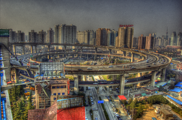 Eine Stadtansicht mit einer zentralen Ringstraße mit Fahrzeugen, einer unteren Straße mit Autos und Fahrrädern, zahlreichen Hochhäusern im Hintergrund, einem Himmel oben, Bäumen auf der linken unteren Seite und kleinen Häusern, die über die Stadt verteilt sind.