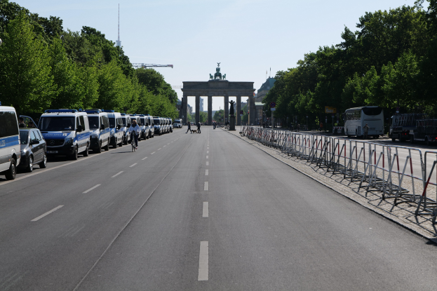 Lange Reihe von Polizeiwagen auf der Seite einer Straße vor dem Brandenburg-Tor in Berlin geparkt, mit Menschen auf Fahrrädern und auf der Straße stehend, Barrieren, Bäume, ein Bogen mit Statuen im Hintergrund und sichtbarer Himmel darüber.