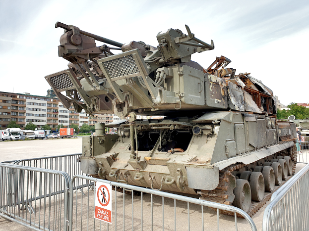 Ein gepanzertes Fahrzeug vor einem Metallzaun mit einem daran befestigten Schild, mit Fahrzeugen, Bäumen, Gebäuden und einem klaren blauen Himmel im Hintergrund.