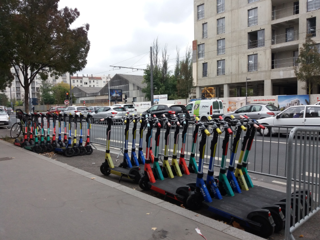 Eine Reihe von E-Scootern, die entlang einer Straße mit einem Geländer, Gebäuden, Bäumen, Pfählen, Verkehrszeichen und Schildern im Hintergrund unter einem klaren blauen Himmel geparkt sind.