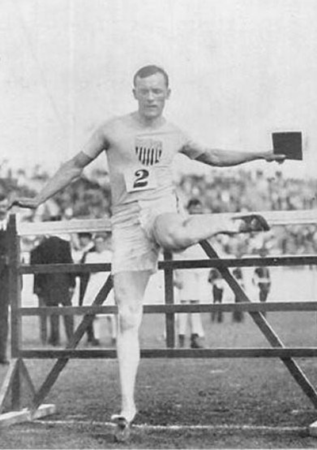 Schwarz-weiß-Foto eines Mannes in der Mitte eines Hindernisrennens, der eine nummerierte Weste und Shorts trägt, mit Zuschauern im Hintergrund und Himmel darüber.