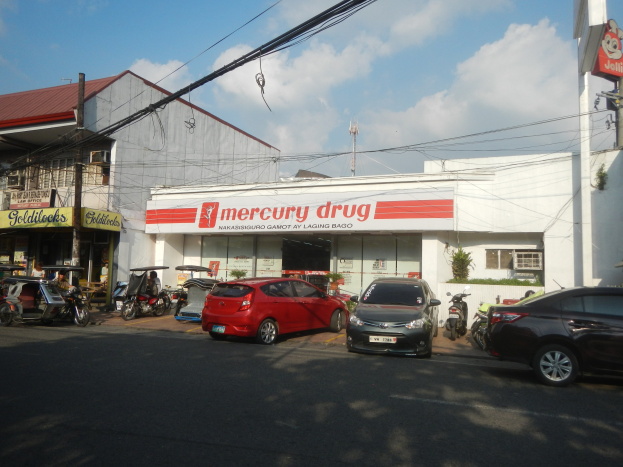 Eine belebte Straßenszene mit Fahrzeugen, Gebäuden mit Schildern, Strommasten mit Drähten und einem bewölkten Himmel, mit einer Mercury Drug Store und parkenden Autos im Vordergrund.
