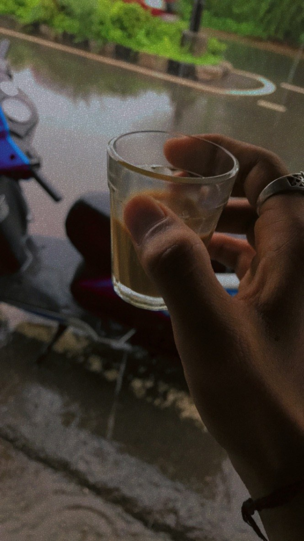 Eine Person mit einem Glas Bier vor einem Fenster, daneben ein Motorroller und im Hintergrund Bäume, Pfähle und eine Straße.