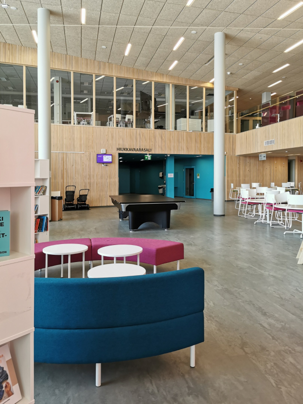 Ein großer Schulinnenraum mit mehreren Tischen, Stühlen, Sofas, Hockern, Bücherregalen, Säulen, Deckenleuchten und einem Wandfernseher.