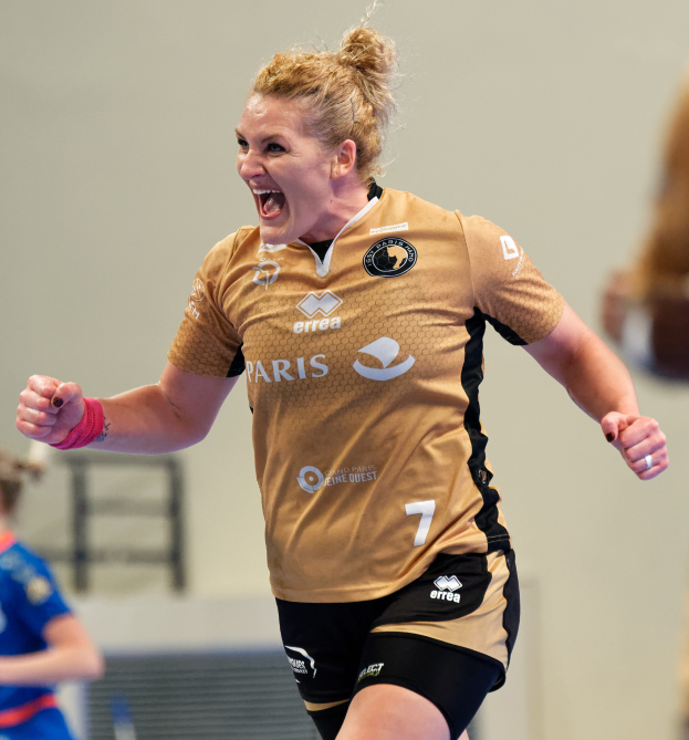 Eine Frau in braunem T-Shirt und schwarzen Shorts feiert nach einem Tor auf einem Futsal-Platz, mit zwei anderen Frauen im Hintergrund.