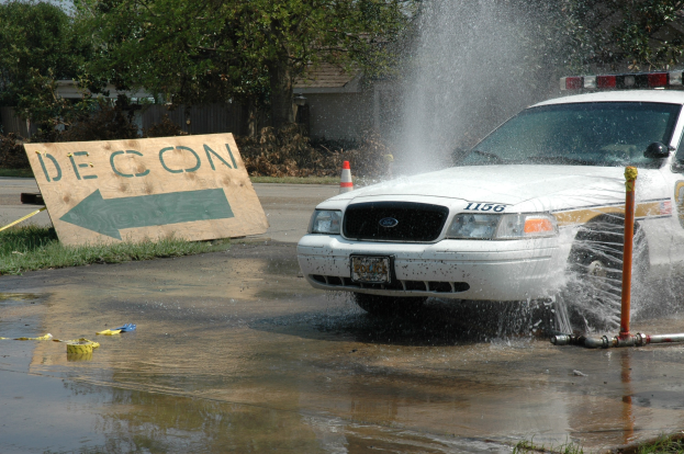 Ein Streifenwagen fährt durch eine Pfütze mit verstreuten Gegenständen auf dem Boden, darunter eine Tafel mit Text, ein Verkehrskegel, Bäume, Gebäude und einen klaren blauen Himmel im Hintergrund.