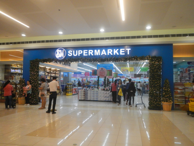 Menschen vor einem Supermarkt, einige halten Einkaufstaschen, mit einem Einkaufswagen, Topfpflanze, Regalen, Schildern und Deckenbeleuchtung.