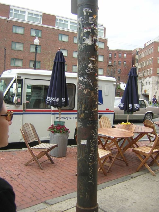 Ein Tisch und Stühle sind auf einem Fußweg aufgestellt, mit Autos und Lieferwagen auf der benachbarten Straße geparkt und Gebäude im Hintergrund sichtbar.