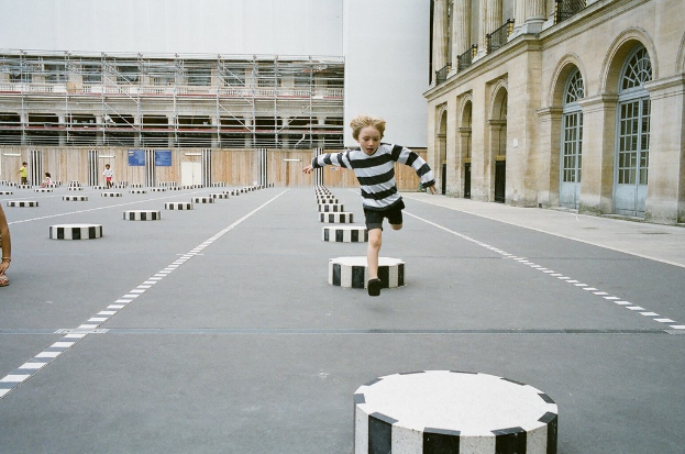 Ein Spielbereich mit vier Kindern, eines läuft auf dem Boden mit schwarzen und weißen Zementstufen, umgeben von einem Schulgebäude.