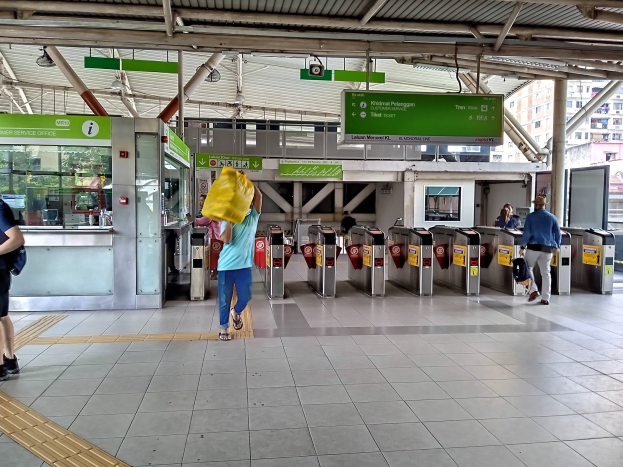 Menschen gehen durch einen Bahnhof mit Metalldetektoren, Hinweisschildern, Metallstangen, Deckenleuchten und Gebäuden mit Fenstern im Hintergrund. Einige tragen Taschen.