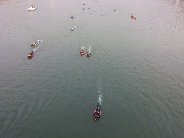 Gruppe von Menschen, die auf einem Gewässer in Kajaks paddeln, mit Bäumen im Hintergrund.