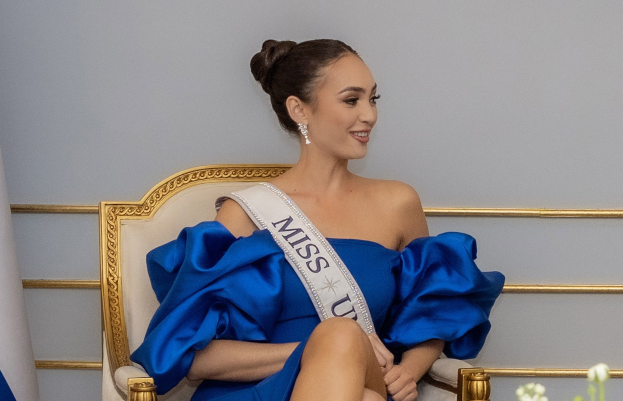 Frau in einem blauen Kleid mit einem "Miss Universe 2019"-Band, die lächelt, während sie auf einem Stuhl sitzt und von Blumen umgeben ist.