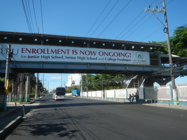 Ein Bus fährt unter einer Brücke mit einem Schild, auf dem "Anmeldung läuft" steht, während Fußgänger darunter gehen und Strommasten die Straße säumen, mit Bäumen und einem klaren Himmel im Hintergrund.