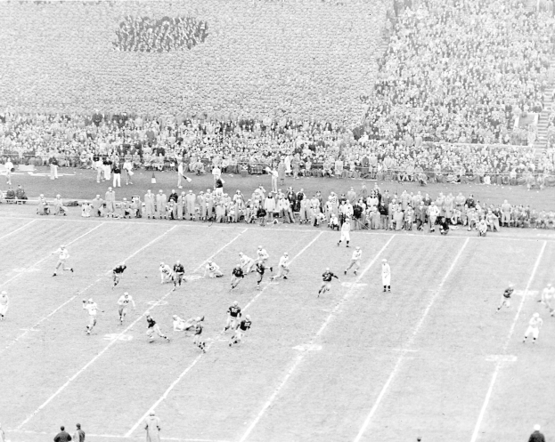 Ein Schwarz-Weiß-Bild eines Fußballspiels in einem Stadion, mit Spielern auf dem Boden und einer großen Zuschauermenge im Hintergrund.