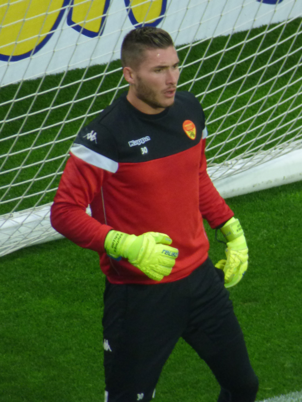 Ein Mann in einem roten Hemd und schwarzen Hosen steht auf einem Fußballfeld mit einem Netz und einer Fahne im Hintergrund, trägt Fußballhandschuhe.