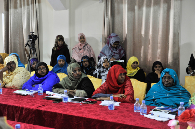 Eine Gruppe von Frauen sitzt an einem Tisch mit Flaschen, Büchern, Mobiltelefonen, Papieren, Stiften und anderen Gegenst├Ąnden und diskutiert mit einer Vase auf der rechten Seite und Vorh├Ąngen im Hintergrund.