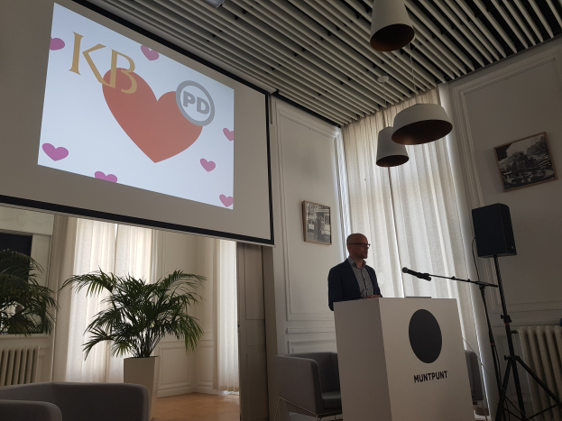 Ein Mann steht an einem Rednerpult mit einem Mikrofon, hält eine Präsentation zum Thema Geschlechtergerechtigkeit, mit einem Projektionsschirm dahinter und einem Raumsetup mit Stühlen, einem Lautsprecher, einer Pflanze, Rahmen, Vorhängen und Deckenleuchten.