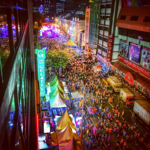 Eine lebhafte Karnevalszene mit einer großen Menge auf der Straße, umgeben von Zelten, Geschäften, Gebäuden und Bannern, mit zahlreichen Lichtern, die den Hintergrundbereich erhellen.