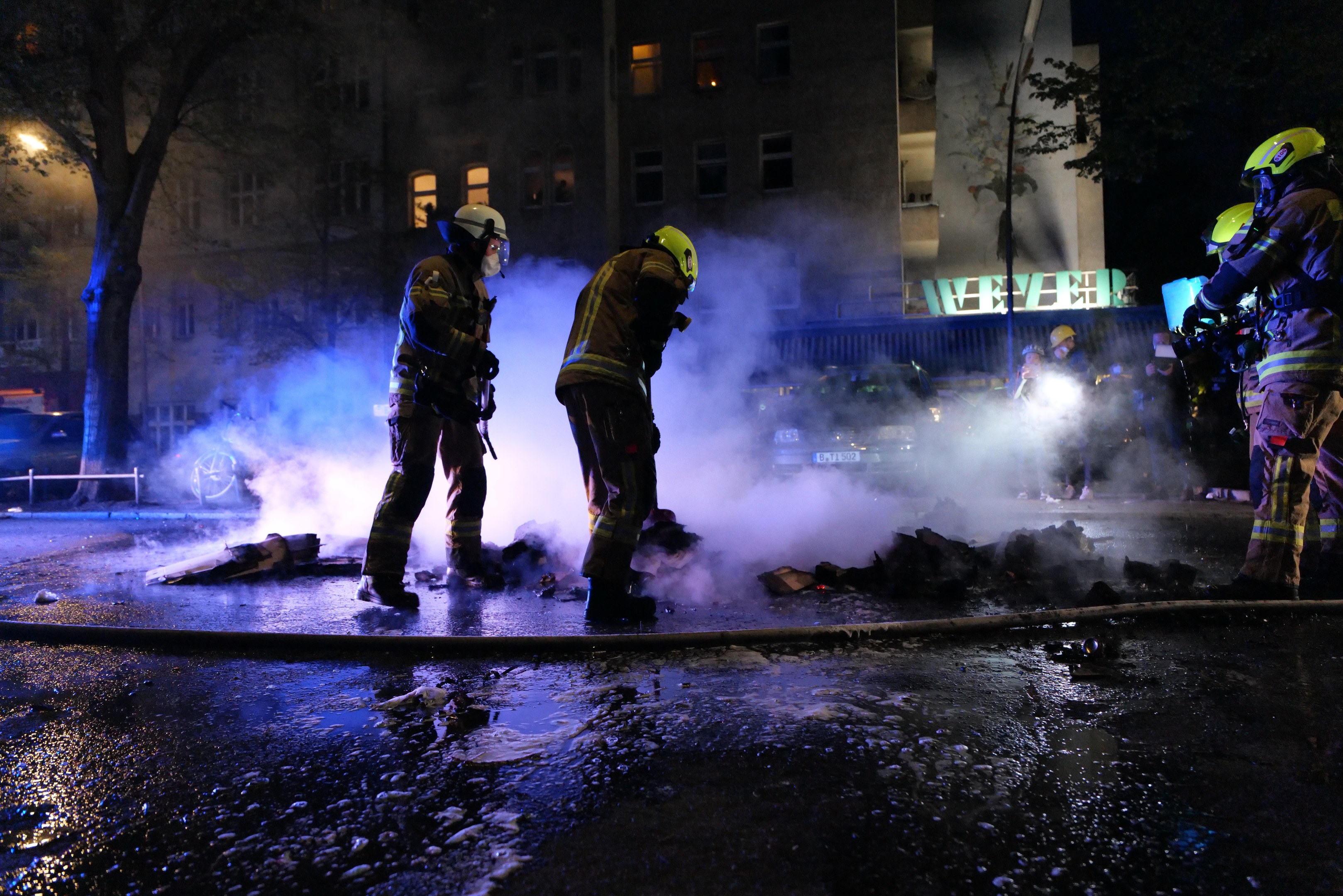 Feuerwehrleute in Helmen stehen nachts auf einer Straße mit Rauch aus einem Rohr, halten Gegenstände; Gebäude, Bäume und Fahrzeuge sind im Hintergrund mit einem Schild auf der rechten Seite sichtbar.