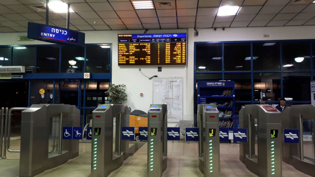 Ein Mann steht vor einer Metalldrehsperre am Eingang eines Flughafens, umgeben von Metalldetektoren, Hinweisschildern, einem Topf mit einer Pflanze, einer Wandtafel, einer Decke mit Leuchten und einem Anzeigebildschirm.