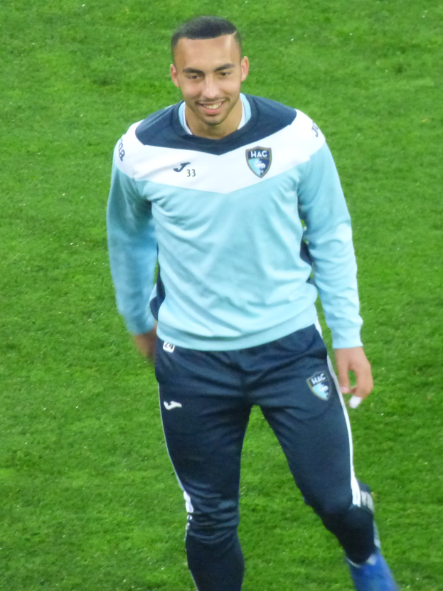 Ein Mann in einem blauen Hemd und schwarzen Hosen lächelt, während er auf einem Fußballfeld geht.