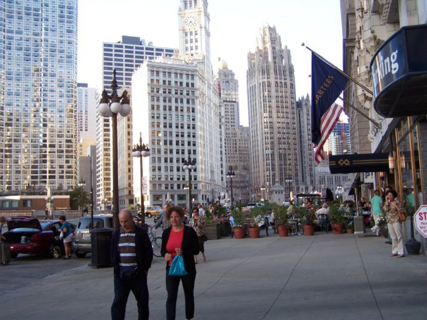 Eine belebte Straßenszene mit Menschen, Blumenkübeln, Autos, Straßenlaternen, Gebäuden, Geschäften und Fahnen auf der rechten Seite.