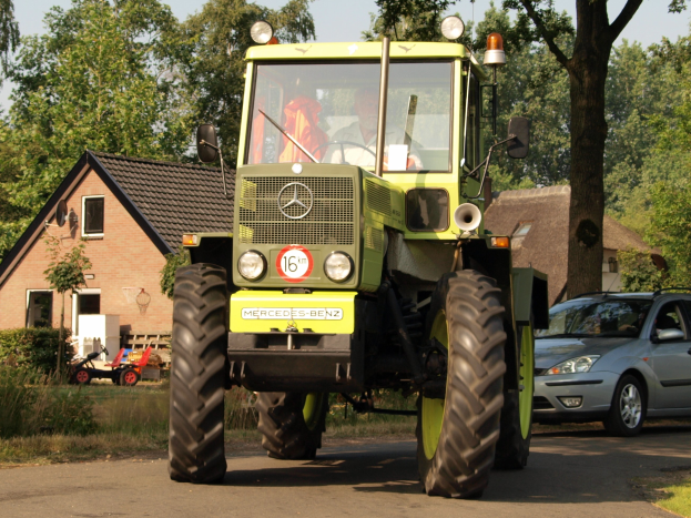 Grüner Traktor auf einer Straße neben einem Auto fahrend, mit einer Person auf dem Fahrersitz des Traktors, vor einer Kulisse aus Häusern, Bäumen, Pflanzen, Gras und einem klaren blauen Himmel.