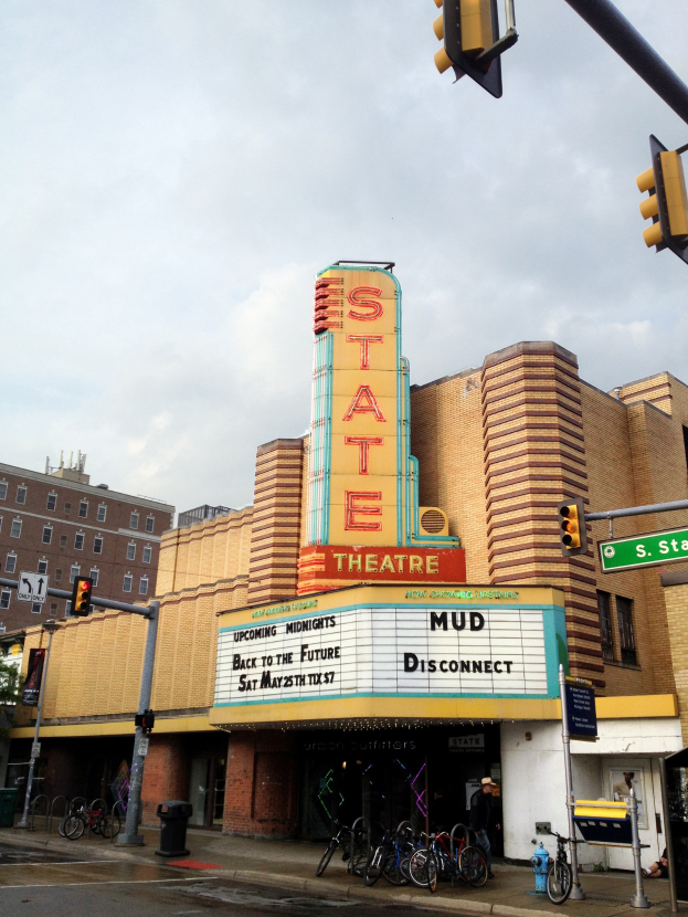 Das Staatstheatergebäude in Detroit, Michigan, mit Fahrrädern auf dem Gehweg, einer Person in der Nähe und umliegenden städtischen Elementen unter einem bewölkten Himmel.