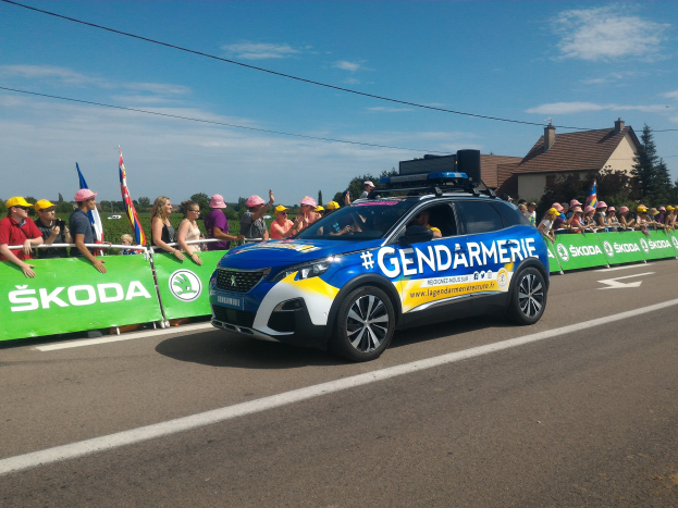 Polizeifahrzeug mit "Gendarmerie de la Tour de France"-Banner an einer Menge mit Fahnen und Mützen tragenden Menschen vorbei, mit Bäumen, Gebäuden und einem bewölktem Himmel im Hintergrund.