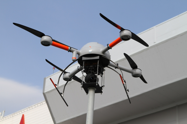 Eine Drohne fliegt über ein Gebäude mit einem Mast im Vordergrund und einem klaren blauen Himmel im Hintergrund.