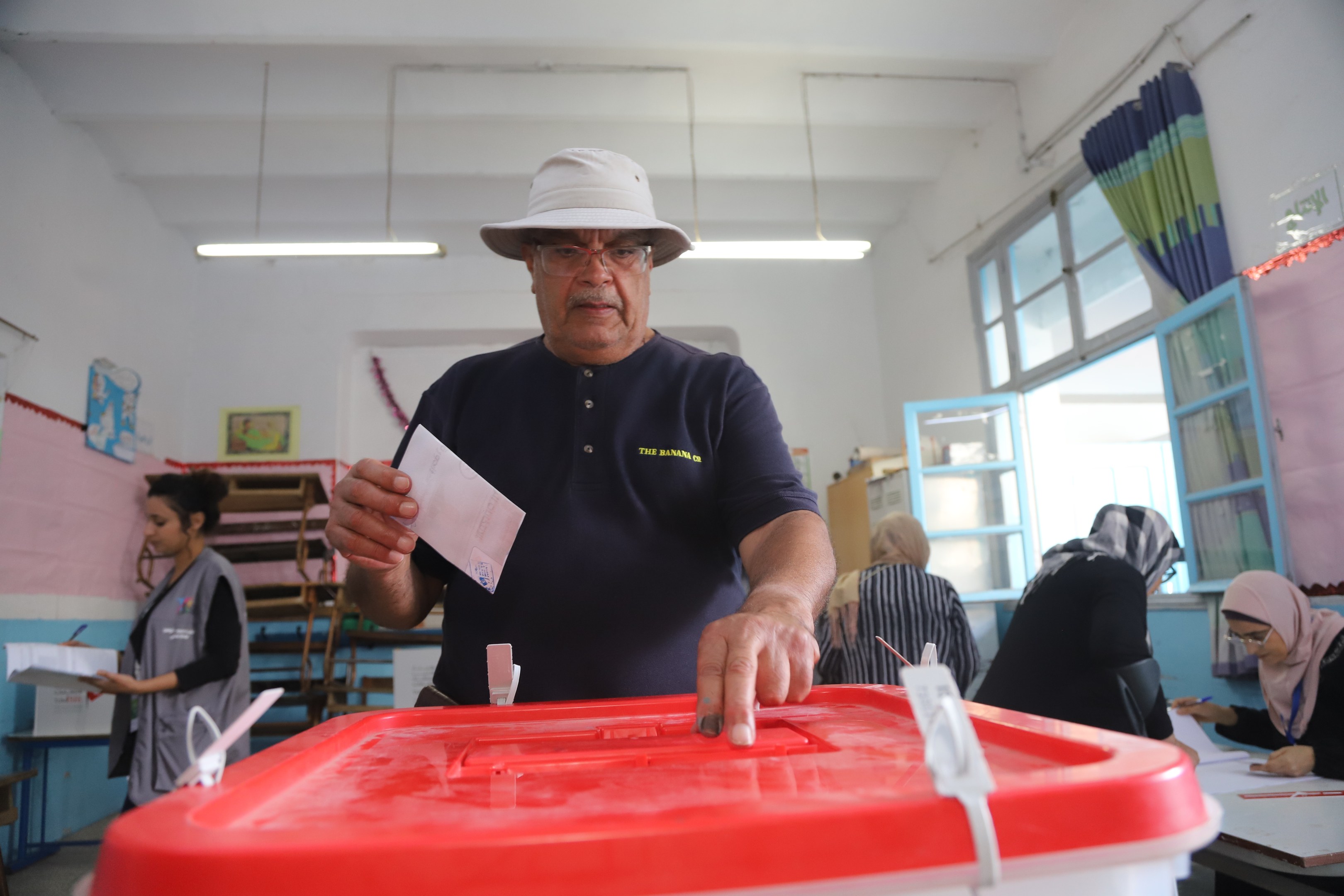 Ein Mann mit Hut wirft seine Stimme, indem er einen Zettel in eine rote Wahlurne steckt, während andere sitzen und im Hintergrund an einem Fenster mit Gardine schreiben.