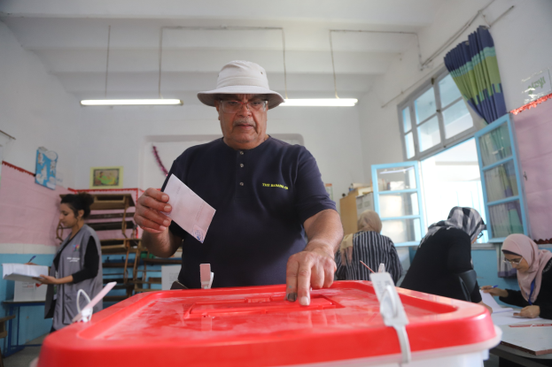 Ein Mann mit Hut wirft seine Stimme, indem er einen Zettel in eine rote Wahlurne steckt, während andere sitzen und im Hintergrund an einem Fenster mit Gardine schreiben.
