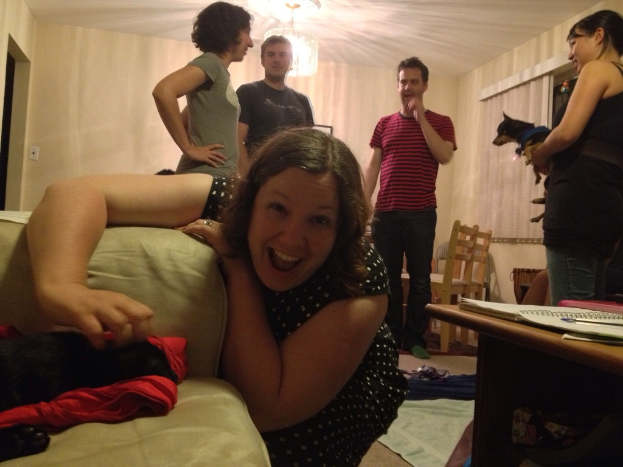 Eine Gruppe von fünf Personen, drei Frauen und zwei Männer, in einem hell erleuchteten Raum mit einem Kronleuchter; eine Frau beugt sich über ein Sofa und lacht, eine andere hält einen Hund und trägt eine schwarze Bluse und Jeans, die dritte Frau hat kurze Haare und helle Haut; die beiden Männer sprechen mit der Frau mit kurzen Haaren; es gibt zwei Stühle in der Ecke und einen Tisch auf der rechten Seite des Raums.