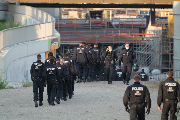 Eine Gruppe von Polizisten geht auf einem Schotterweg, mit einer Wand auf der linken Seite und Pflanzen auf der rechten Seite, mit einer Brücke und anderen Gegenständen im Hintergrund.