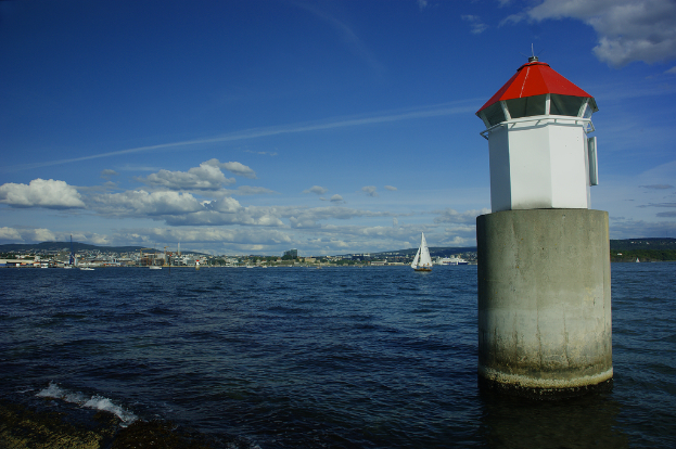 Boote auf dem Meer mit einem Leuchtturm im Vordergrund, Gebäude, Berge und der Himmel im Hintergrund.