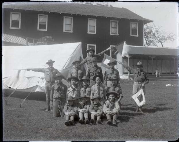 Gruppe von Pfadfindern in Uniformen und Hüten, die vor einem Zelt für ein Schwarz-Weiß-Foto posieren, wobei einige auf dem Gras sitzen und andere stehen; im Hintergrund ein Gebäude, Bäume und ein klarer Himmel sichtbar.