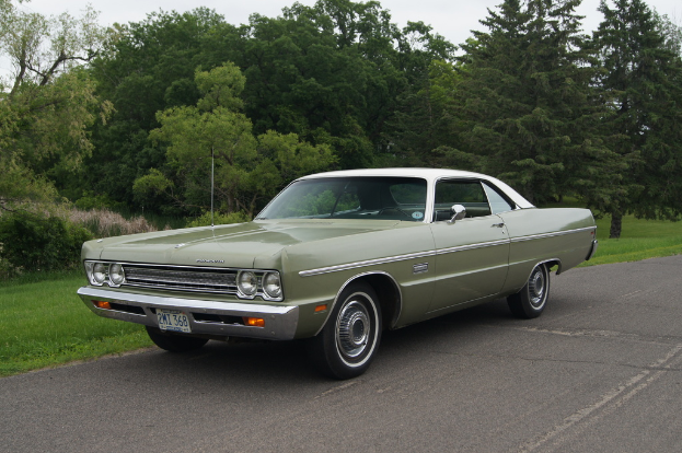 Ein grünes Oldtimer-Auto fährt auf einer Straße mit Bäumen und Wiesen dahinter unter einem klaren Himmel.