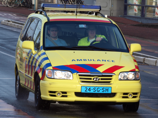 Ein gelber Krankenwagen mit zwei Insassen fährt an parkenden Fahrrädern auf einem Gehweg vorbei, mit einem Glasfenster-Gebäude im Hintergrund.