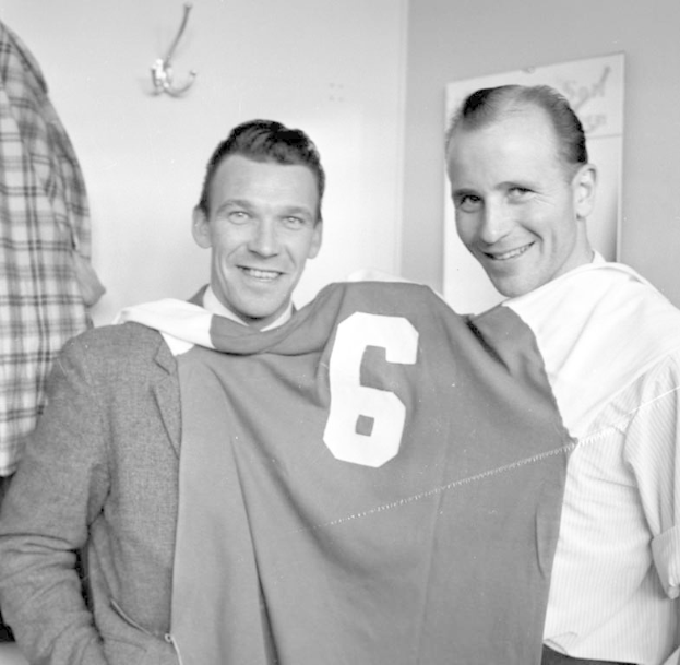 Zwei Männer in Schwarz-Weiß, die strahlend ein Fußballtrikot hochhalten, mit einer Tafel und einem Kleiderbügel an der Wand dahinter.