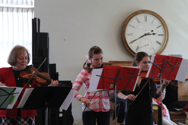 Drei Frauen auf der Bühne mit Geigen stehend und Musiknotenständer vor ihnen, ein Publikum rechts sitzend, eine Uhr an der weißen Wand dahinter und ein weißer Vorhang in der linken Ecke.
