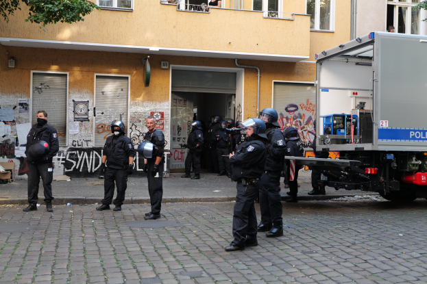 Eine Gruppe von Polizisten in Helmen und mit Waffen steht vor einem Gebäude mit Fenstern, Rollläden und Graffiti, neben einem parkenden Lkw und Bäumen im Hintergrund.