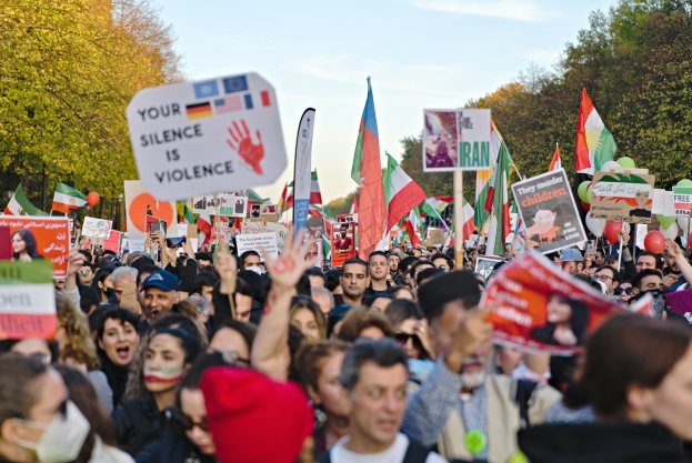 Eine große Gruppe von Menschen mit Schildern und Flaggen im Zusammenhang mit der iranischen Revolution, mit Bäumen im Hintergrund und einem klaren blauen Himmel darüber.
