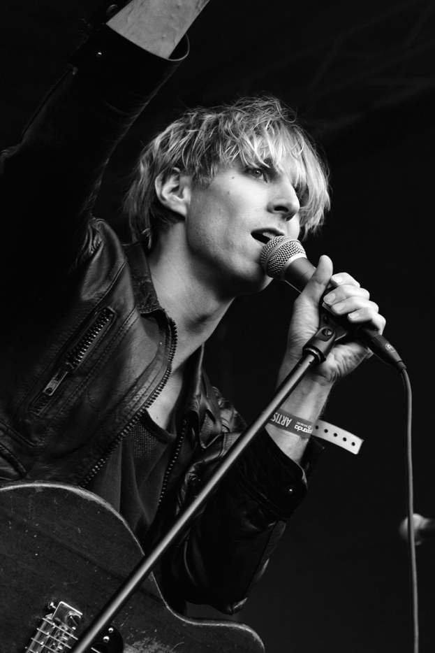 Schwarzes und weißes Foto eines Mannes in einer Lederjacke, der in ein Mikrofon singt, während er auf der Bühne eine E-Gitarre spielt.