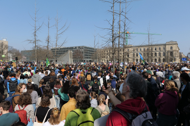 Eine große Menge Menschen steht vor einem Gebäude mit Fenstern, Bäumen und einem klaren Himmel im Hintergrund, viele halten Schilder und tragen Taschen, was auf eine Klimawandel-Demonstration hindeutet.