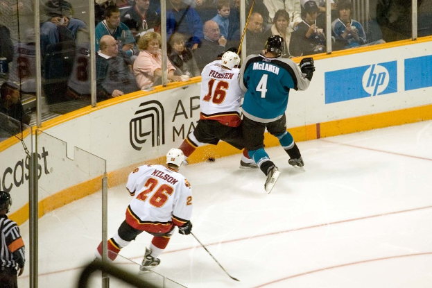 Ein Mann in einem sechzehn Nummern Trikot, Helm, Shorts und Schuhen steht neben einem anderen Mann in einer blauen Jacke, Brille, Helm und Hose neben einem Fenster, mit einem Publikum, das oben sitzt und ein Spiel beobachtet, einem Mann in einem vierundzwanzig Nummern Trikot, der unten einen Stock hält, einem Schiedsrichter hinter Glas in der unteren linken Ecke und einem Werbeplakat auf der rechten Seite.