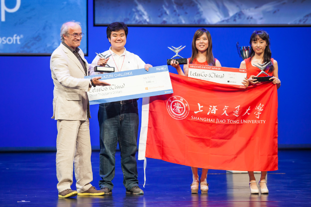 Vier Personen auf einer Bühne mit Auszeichnungen, Schecks und einer Fahne, die fröhlich lächeln und ein digitaler Bildschirm mit einer Projektion im Hintergrund.