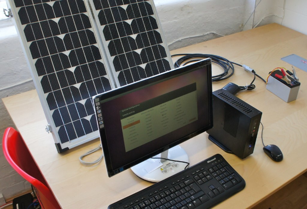 Ein Computersystem mit Tastatur, Maus und CPU auf einem Tisch, neben einem Solarpanel, einer Batterie, Chips und Kabeln, mit einem roten Stuhl und einer weißen Wand im Hintergrund.